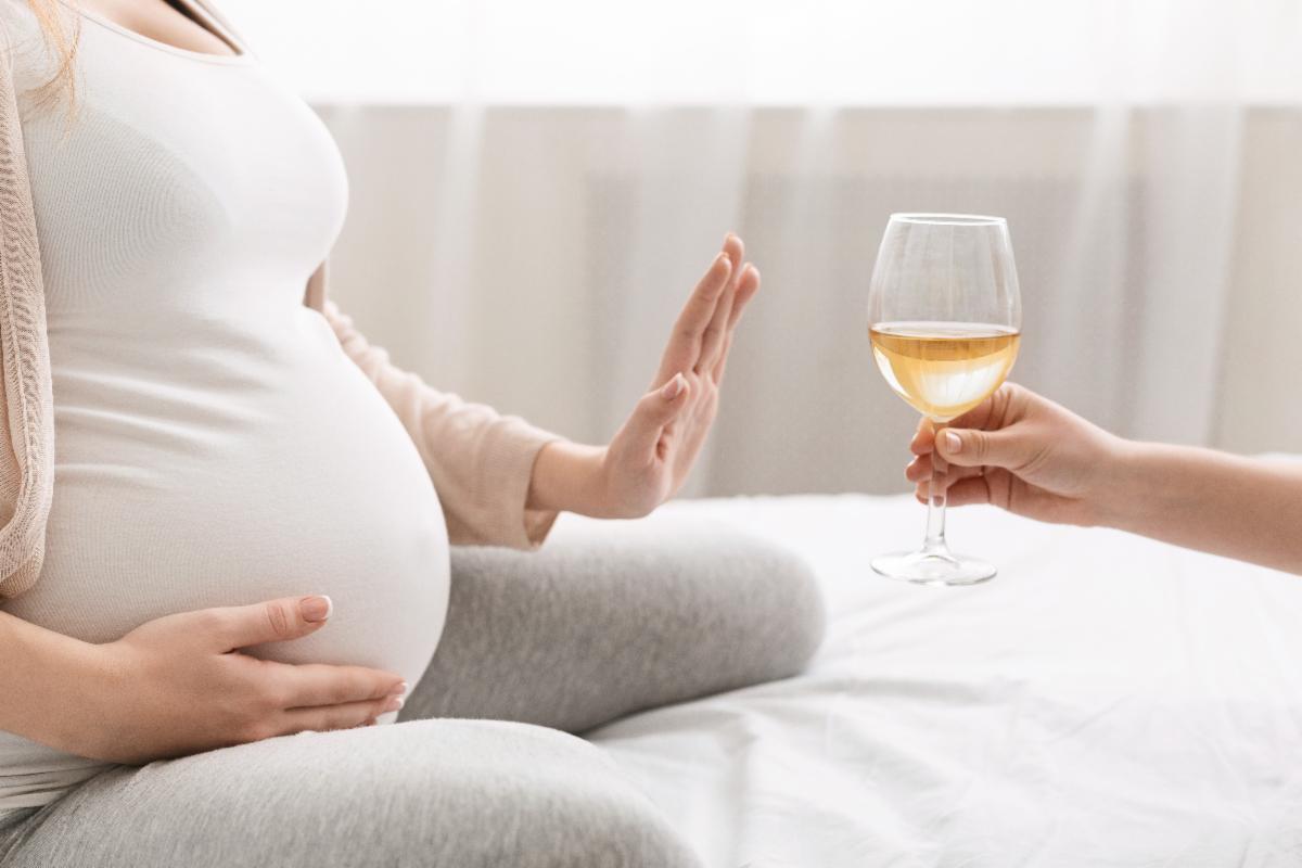A pregnant person sitting on a bed refusing a glass of wine.