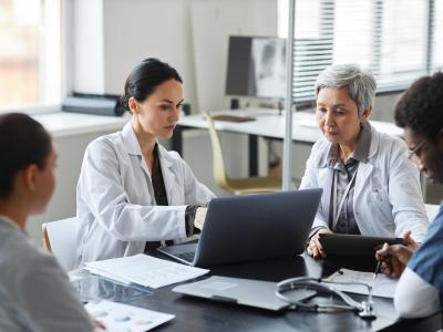 Brunette female clinician in lab coat pointing at laptop screen while making presentation of medical data to group of intercultural colleagues