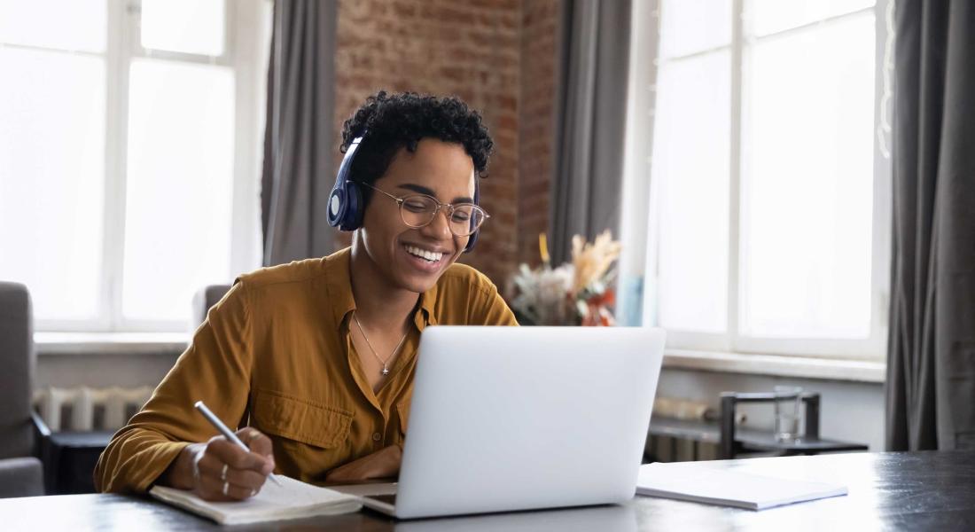 smiling Black woman wearing headphones looking at her laptop and taking notes