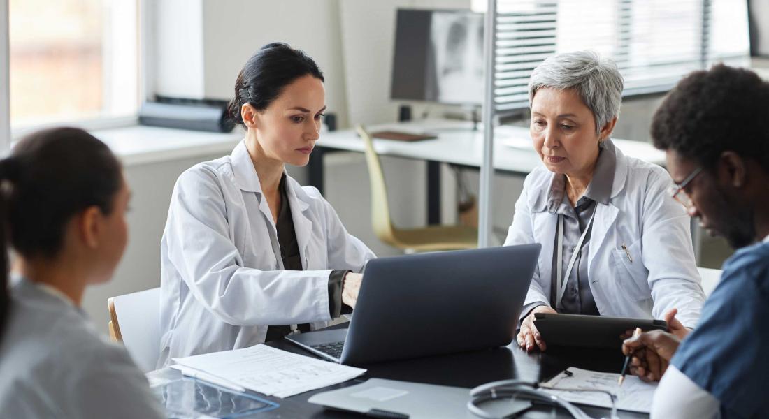 Brunette female clinician in lab coat pointing at laptop screen while making presentation of medical data to group of intercultural colleagues 