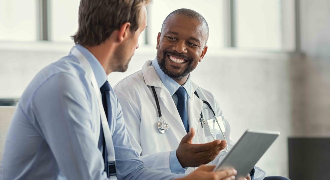 Black man in white coat talking to white man holding a tablet